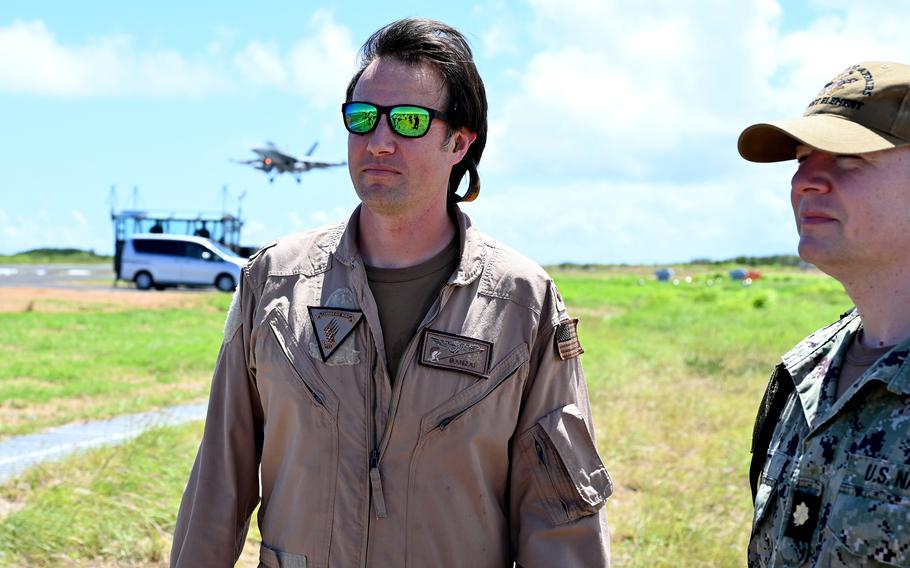 A man in a tan flight suit and sunglasses stands in a grassy area as a military fighter jet lands in the background.
