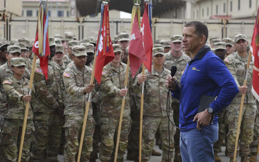 Michael Grinston talks into a microphone to a group of U.S. soldiers, some holding flags.
