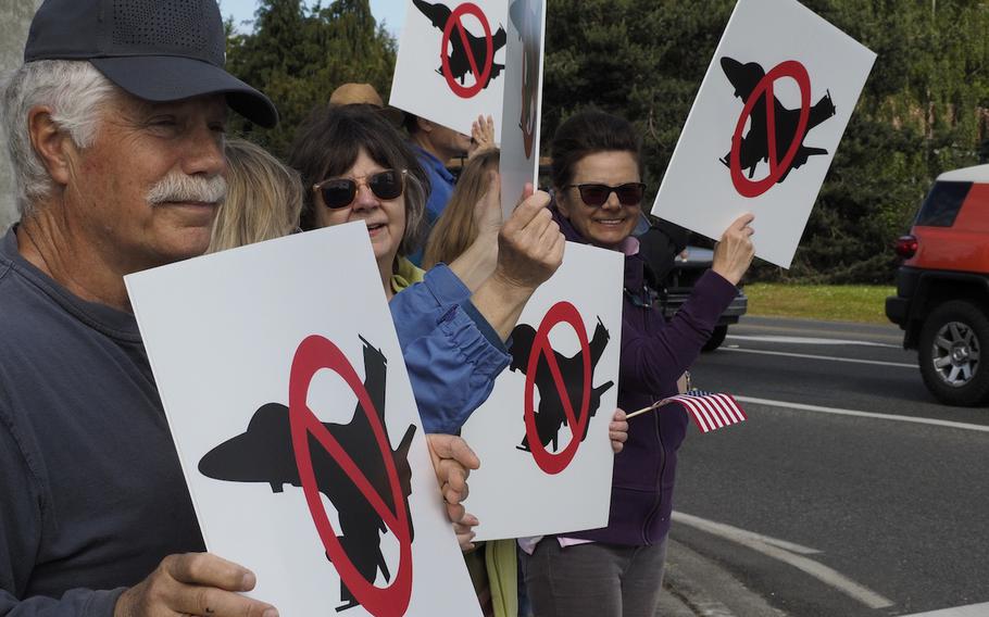 Protesters stand on a sidewalk holding up signs with a black outline of a fighter jet with a red cross over it.