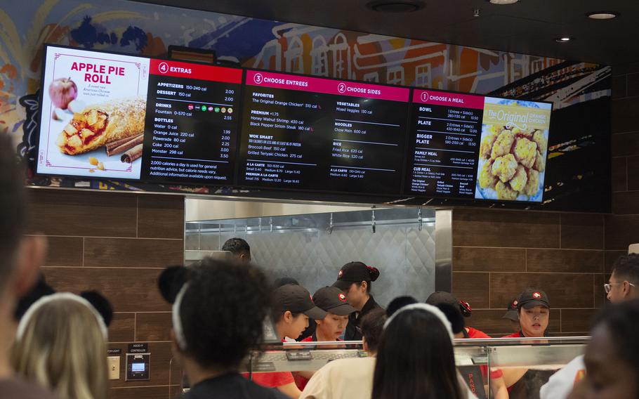 Panda Express restaurant interior showing digital menu boards above the serving counter. Kitchen staff in black caps work behind the counter while customers wait to order.