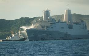 The USS New Orleans amphibious transport dock receives water sprays from a Japan Self-Defense Force vessel and two tugboats near White Beach Naval Facility in Okinawa. The large gray warship with hull number 18 is anchored in blue waters with forested hills visible in the background. 