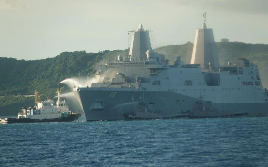 The USS New Orleans amphibious transport dock receives water sprays from a Japan Self-Defense Force vessel and two tugboats near White Beach Naval Facility in Okinawa. The large gray warship with hull number 18 is anchored in blue waters with forested hills visible in the background. 