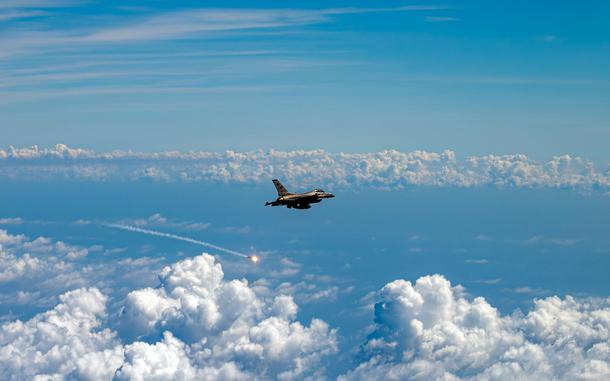 A military fighter jet flying high above the clouds against a blue sky, with a visible projectile trail streaking away from the aircraft.