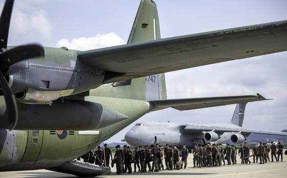 Military personnel in camouflage uniforms line up on an airport tarmac next to a large green military transport aircraft with its rear cargo ramp lowered. 