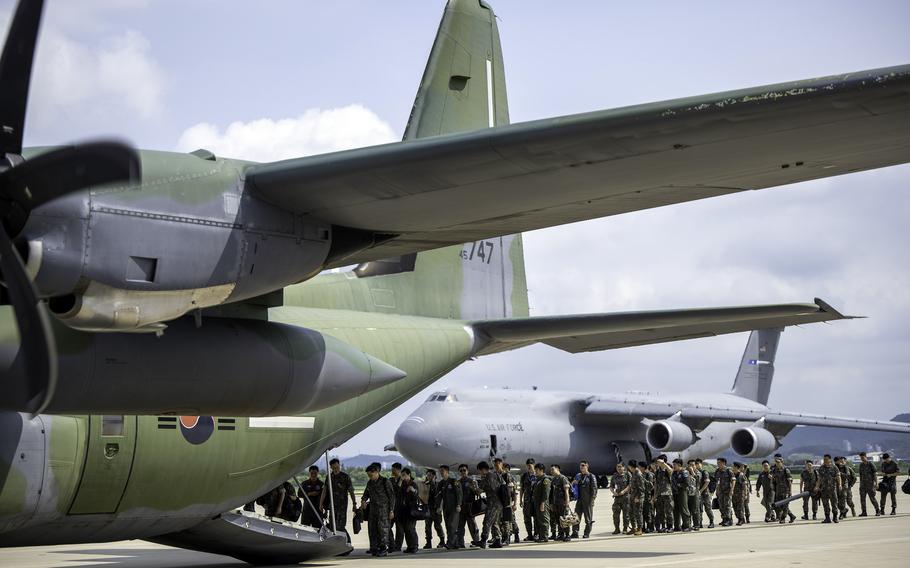 Military personnel in camouflage uniforms line up on an airport tarmac next to a large green military transport aircraft with its rear cargo ramp lowered. 