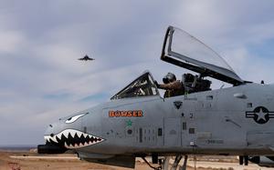 A gray U.S. Air Force A-10C Thunderbolt II sits on the tarmac as its pilot, wearing full flight gear, runs a systems check. The fighter jet displays the USAF emblem and its nose is painted with eyes and a mouth with razor sharo teeth. The name “Bowser” is painted in oragne with a teal star underneath. The desert background is interrupted by another jet in the sky.