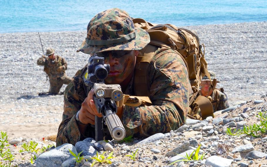 A Marine in camouflage combat gear aims a rifle while lying on his stomach atop a dune of rocks and sand, as another Marine in camouflage combat gear kneels on a beach in the background.