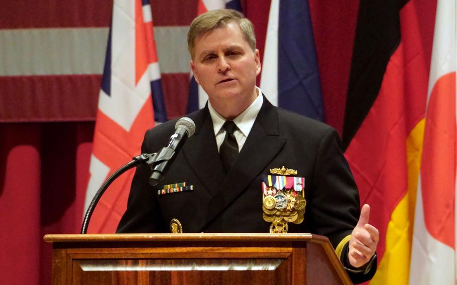 A naval officer in black dress uniform stands at a wooden podium and speaks into a microphone in front of a row of international flags.