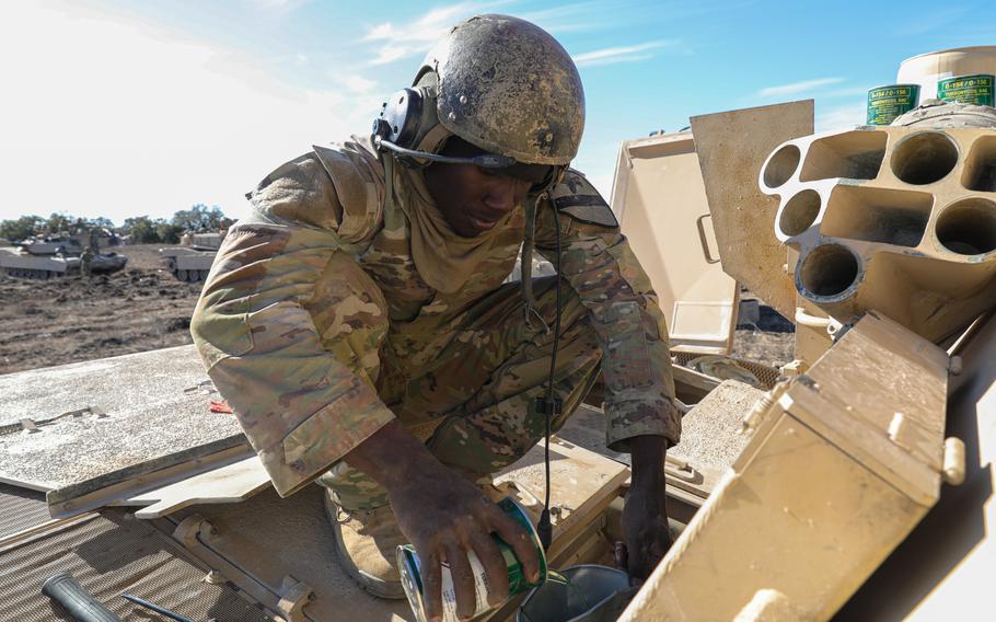 A soldier in camouflage uniform and helmet crouches down while atop a tank and pours liquid from a can into a funnel.