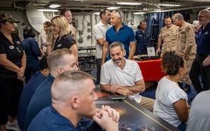 A group of military personnel and civilians gather around a table in a naval vessel. The room features typical naval interior elements including exposed pipes, fluorescent lighting and blue carpeting.
