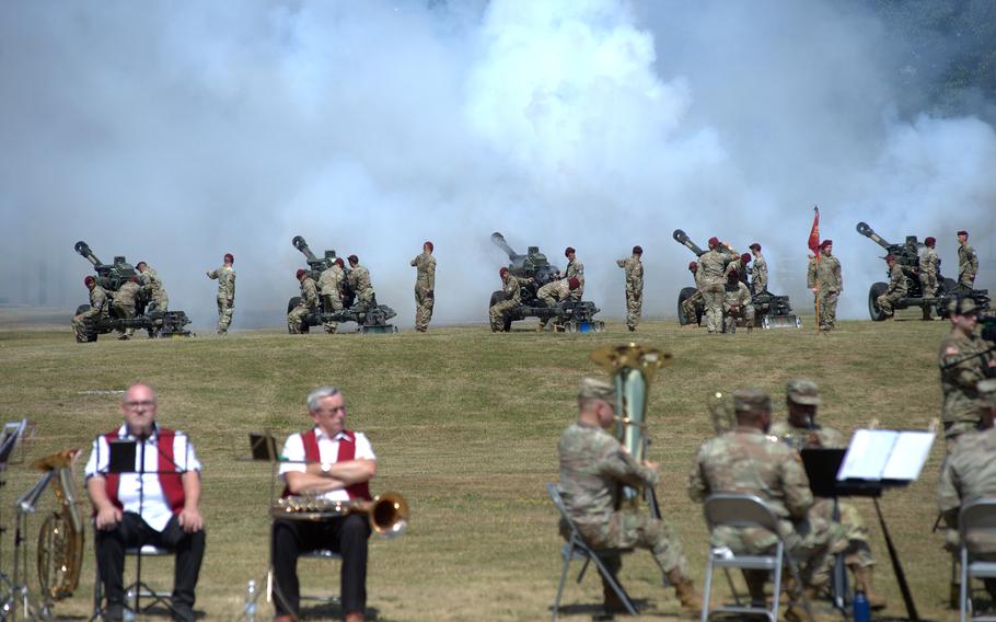 The band plays in front, while smoke rises in the background as artillery fires.