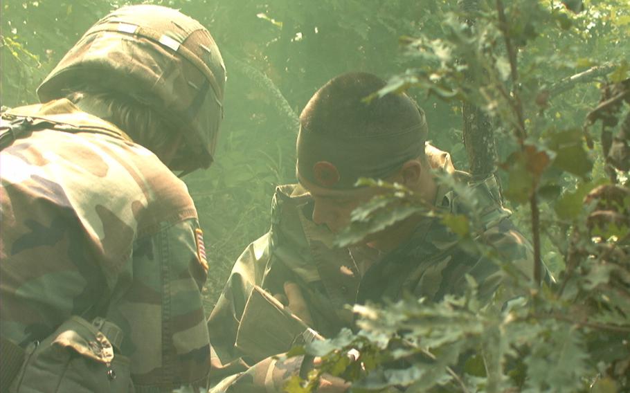 Two soldiers in camoflauge are seen from the shoulders up, partially obscured by leaves.
