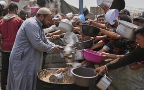 A row of Palestinian children hold out pots and bowls to receive food while a man in a long robe pours food into one.