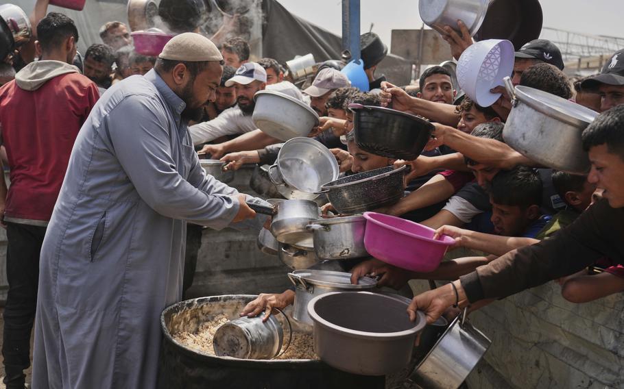 A row of Palestinian children hold out pots and bowls to receive food while a man in a long robe pours food into one.