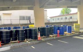 Black and blue industrial barrels are lined up on wooden pallets under a concrete structure, with orange traffic cones and a fence in the background.