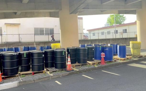 Black and blue industrial barrels are lined up on wooden pallets under a concrete structure, with orange traffic cones and a fence in the background.