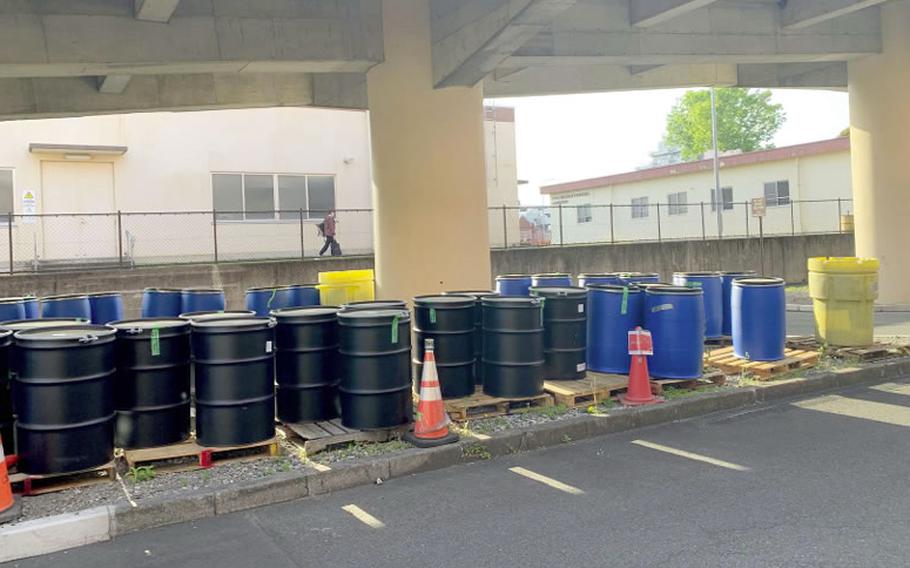 Black and blue industrial barrels are lined up on wooden pallets under a concrete structure, with orange traffic cones and a fence in the background.