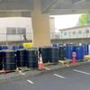 Black and blue industrial barrels are lined up on wooden pallets under a concrete structure, with orange traffic cones and a fence in the background.