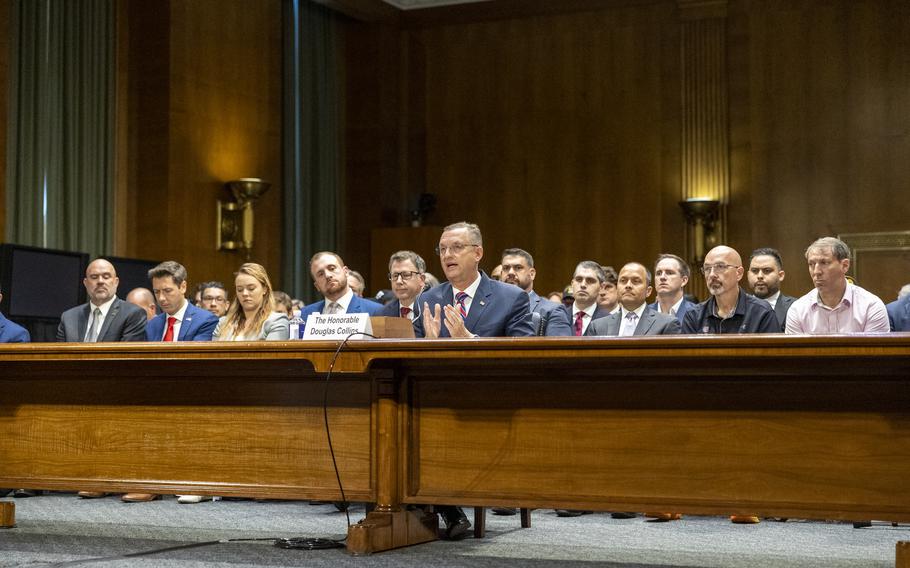 Doug Collins gestures in the middle of a long table as rows of people watch behind him.