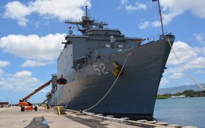 A naval ship, seen from ground level, sits in water moored to a concrete pier with clouds and bluesky behind it.