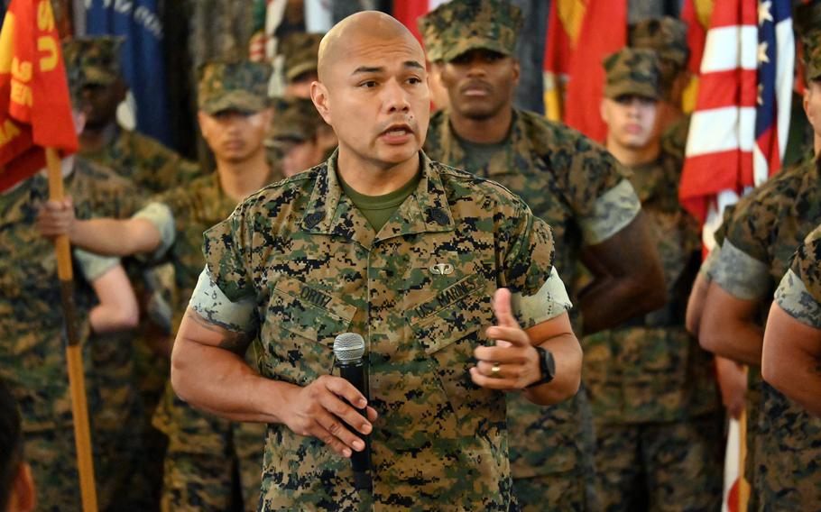 A Marine in camouflage uniform holds a microphone in his right hand while gesturing with his left hand as he speaks in front of other Marines in camouflage uniforms standing at attention.