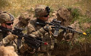 Two soldiers in full combat gear and camouflage uniforms are positioned in a trench. The terrain around them shows disturbed earth and vegetation typical of a training or operational environment.
