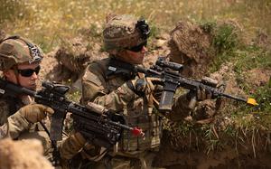 Two soldiers in full combat gear and camouflage uniforms are positioned in a trench. The terrain around them shows disturbed earth and vegetation typical of a training or operational environment.
