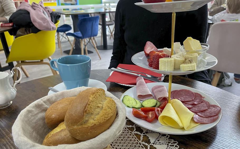 A tiered serving platter holds cold cuts, fruit, and jams next to a bread basket and coffee.
