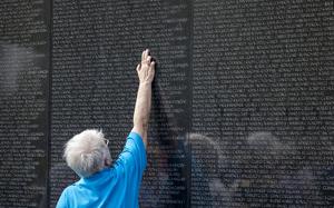 A man in a blue shirt reaches up with his arm to touch a name engraved on a black memorial wall.