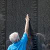 A man in a blue shirt reaches up with his arm to touch a name engraved on a black memorial wall.