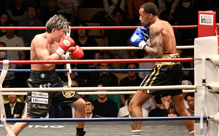 Two boxers in black shorts, one wearing blue gloves and the other wearing red gloves, face each other in a ring with a crowd in the background.