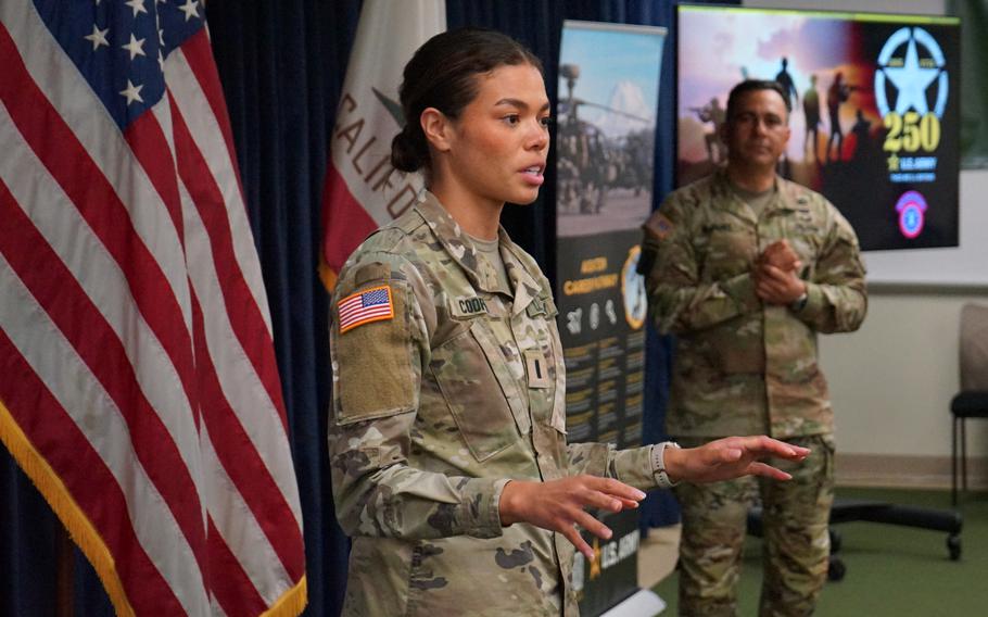 A soldier speaks with her hands out in front of the USA flag