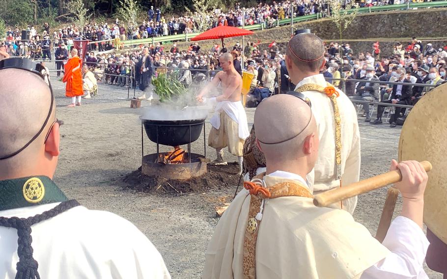 A shirtless Japanese monk stands before a cauldron over an open flame as other monks and a crowd of spectators watch.