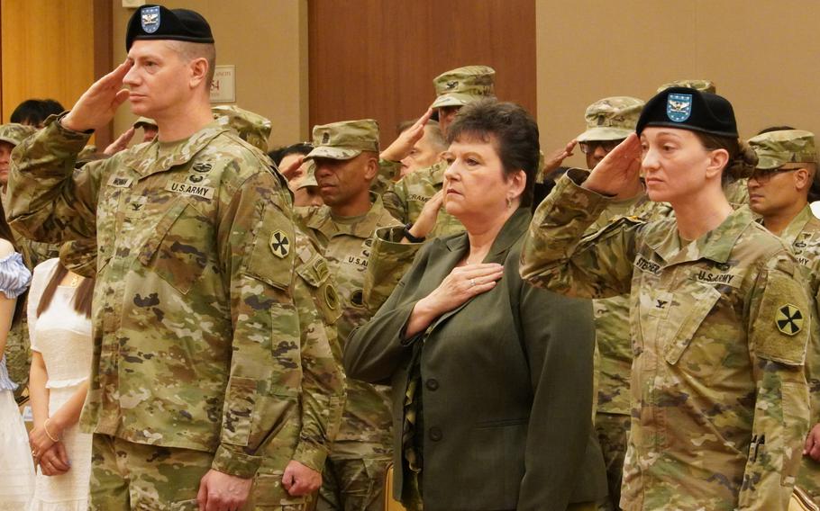 A group of U.S. Army soldiers stand at attention. A civilian woman in a dark green business suit stands among them with her hand over her heart. 