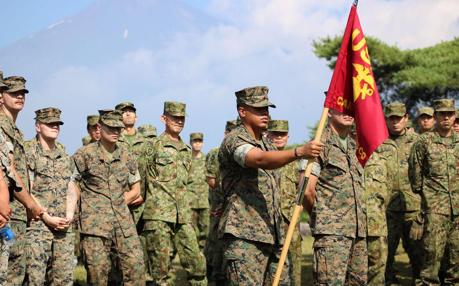 A formation of U.S. and Japanese service members in various camouflage uniforms stands at attention outdoors; one Marine holds a red unit guidon with “USMC” and “Camp” partially visible, with Mount Fuji faintly visible in the background.