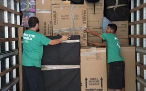 Two moves in green shirts and black shorts pack cardboard boxes into stacks in the back of a truck.