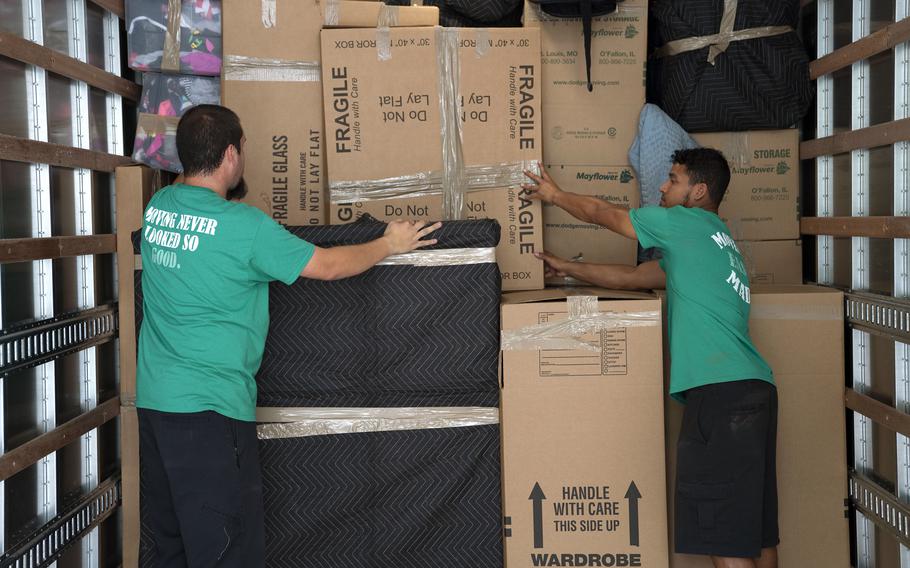 Two moves in green shirts and black shorts pack cardboard boxes into stacks in the back of a truck.