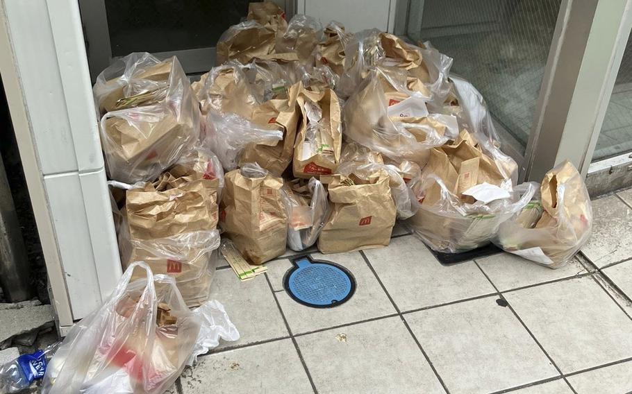 A large pile of McDonald’s takeout bags and plastic bags stacked on a tiled floor outside glass doors. The brown paper bags with McDonald’s logos are bundled together in clear plastic bags.