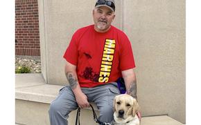 A man wearing a red Marines T-shirt and a ball cap sitting with his dog next to him.