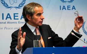 A man in a suit gestures at a podium with the International Atomic Energy Agency logo emblazoned on a poster or wall behind him.