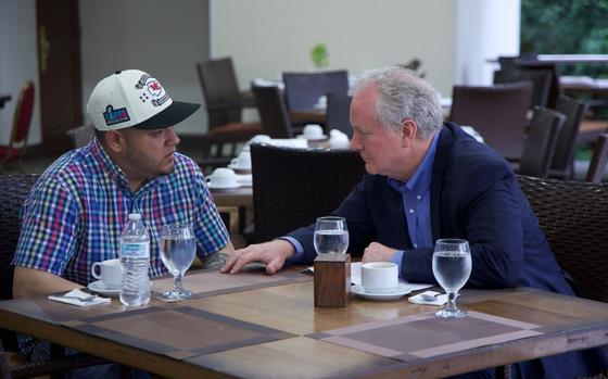 Two men sit across from each other at a table engaged in conversation. One wears a colorful plaid shirt and baseball cap, the other a navy blazer. Cups of coffee and glasses of water rest between them.