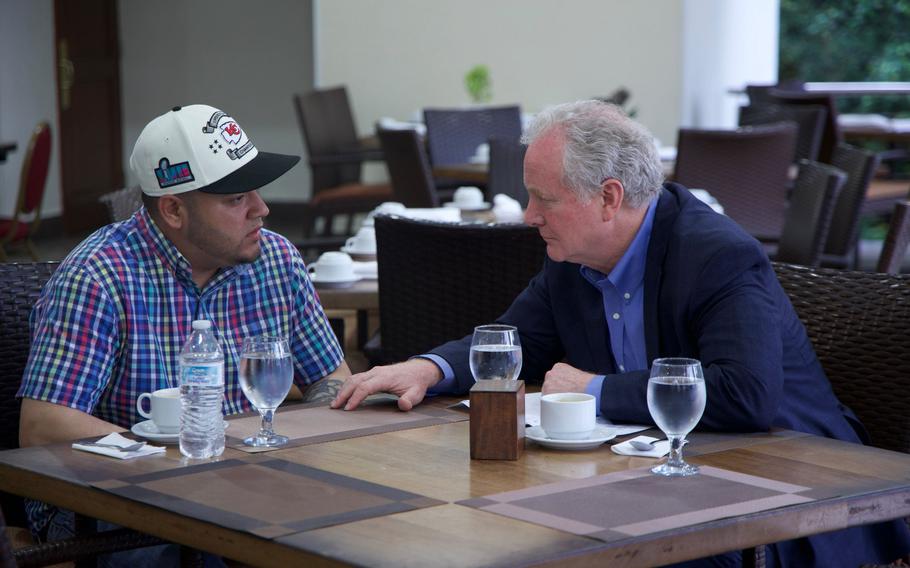 Two men sit across from each other at a table engaged in conversation. One wears a colorful plaid shirt and baseball cap, the other a navy blazer. Cups of coffee and glasses of water rest between them.
