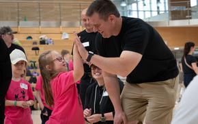 A girl with a pink shirt and glasses gives a high five to a man in a black T-shirt and khaki shorts in a gym at Spangdahlem Air Base.