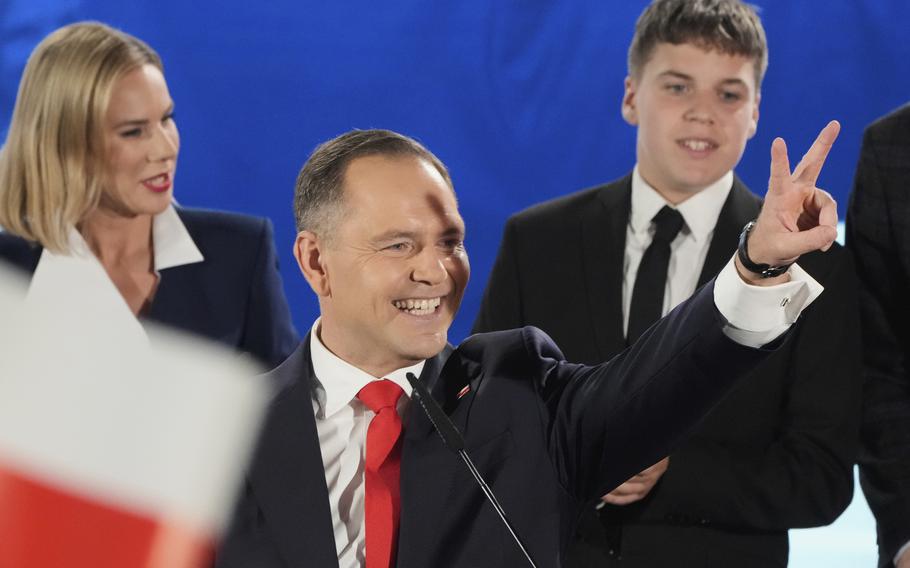 A man in a suit and red tie holds up a V-sign with his fingers. The red and white Polish flag can be seen in the left foreground.