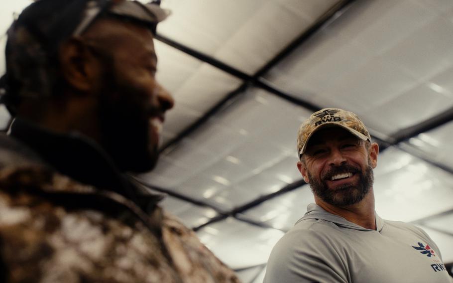 Two men smiling at each other, one out-of-focus near the camera and the other in-focus farther away, on the right.