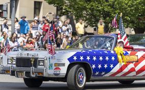 A car with stars and stripes and an American flag rolls down the street.