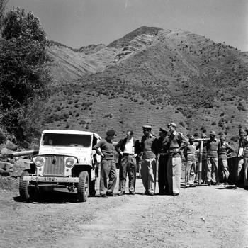 Two groups of men, some standing near a jeep, another group hanging back.