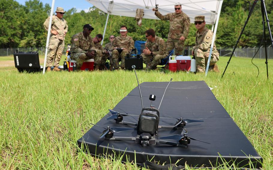 A small drone on a field in the foreground, with soldiers under a tent in the background.