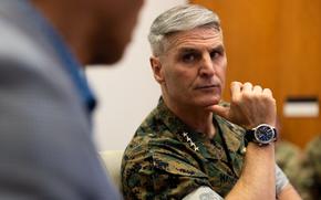 A Marine Corps officer in camouflage uniform sits in a meeting room with his thumb on his chin as he listens to another person speak.