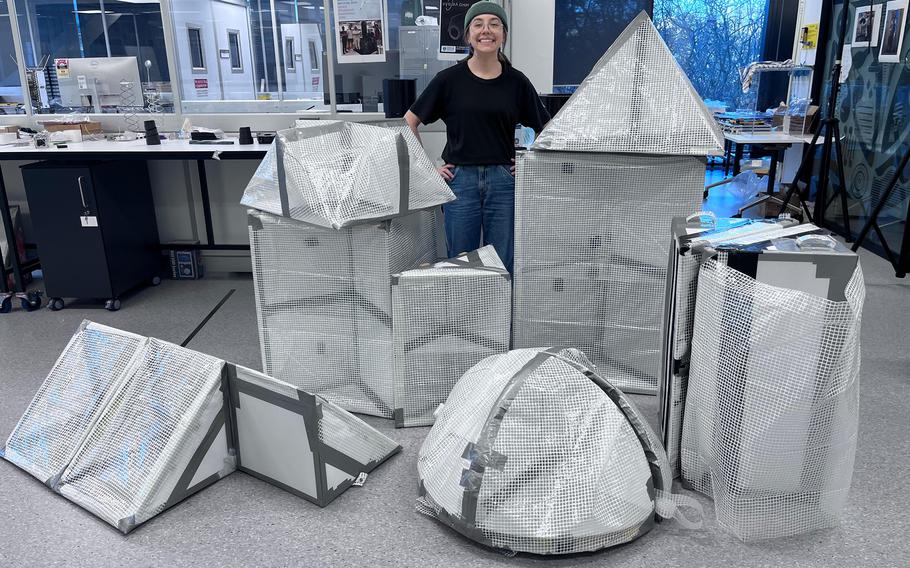 A smiling woman in a black T-shirt and green beanie stands in a laboratory or research facility surrounded by various geometric shapes covered in reflective metallic mesh material. 